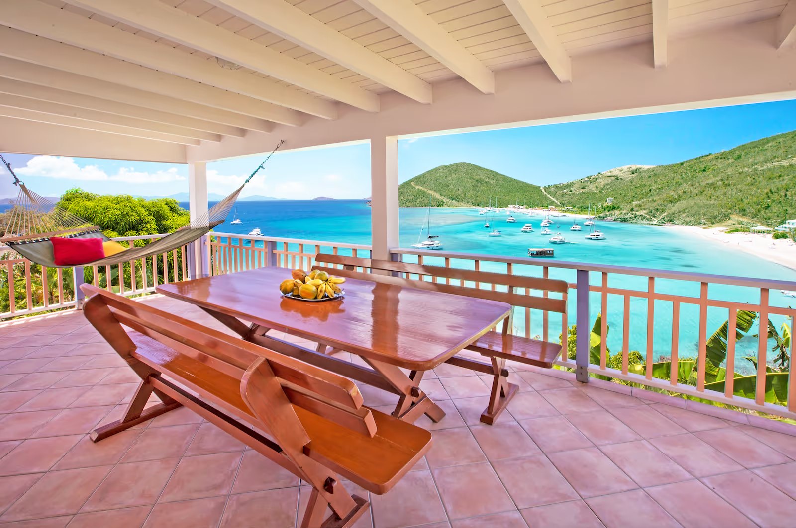 Villa deck at White Bay Villas — wooden bench and table, hammock with colorful cushion, overlooking the turquoise waters of White Bay with sailboats anchored in the bay and green Caribbean hills on the horizon